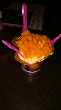 A cup of ice cream topped with fresh mango chunks is presented on a dark wooden table. Three bright pink plastic spoons are inserted into the ice cream, and the treat is placed in a waffle cone bowl within a colorful paper cup.