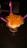 A cup of ice cream topped with fresh mango chunks is presented on a dark wooden table. Three bright pink plastic spoons are inserted into the ice cream, and the treat is placed in a waffle cone bowl within a colorful paper cup.