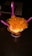 A cup of ice cream topped with fresh mango chunks is presented on a dark wooden table. Three bright pink plastic spoons are inserted into the ice cream, and the treat is placed in a waffle cone bowl within a colorful paper cup.