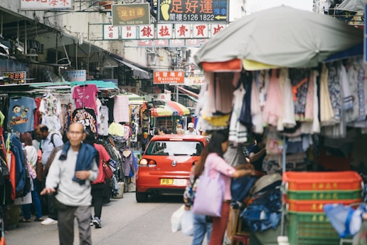 A bustling street market with various stalls selling clothes and other goods. People are walking, shopping, and passing by. A red car is parked in the middle, surrounded by a lively atmosphere and bright, colorful signs with text above the stalls.