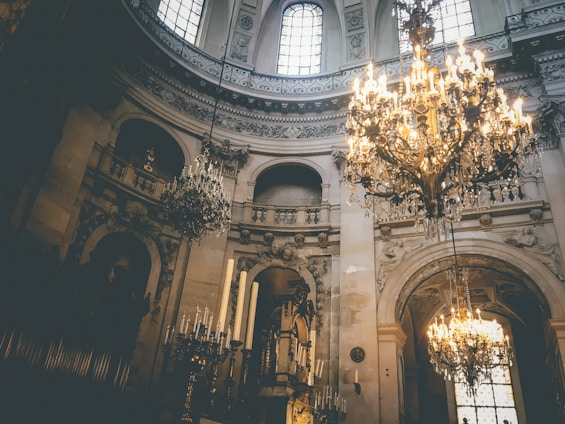 Intricate chandeliers hang from a grand, ornate ceiling in a historical interior space with arched windows and detailed architectural elements. The lighting is warm, casting a golden glow throughout the room, highlighting the elegant decorations and sculptures.