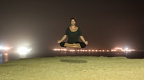 A person is levitating in a seated position with legs crossed and arms relaxed on their knees against a nighttime backdrop. The background features a body of water with glowing lights reflecting off the surface, creating a serene atmosphere.