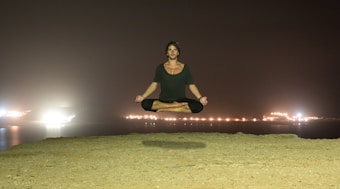 A person is levitating in a seated position with legs crossed and arms relaxed on their knees against a nighttime backdrop. The background features a body of water with glowing lights reflecting off the surface, creating a serene atmosphere.