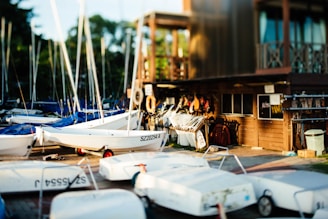 A collection of small sailboats with white hulls and blue tarps are lined up on a dock. The scene includes a wooden building on the right side, which appears to be a boathouse or storage area, with various boating equipment and life jackets visible. The area is well-lit, suggesting a sunny, late afternoon ambiance.