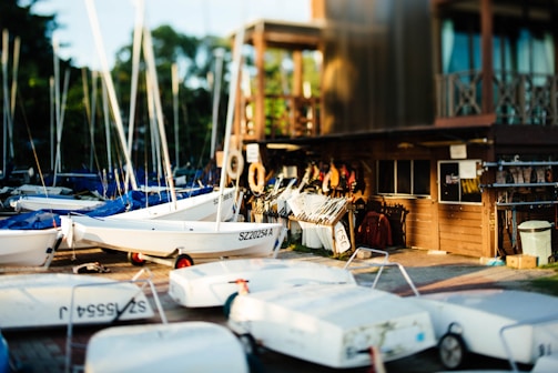 A collection of small sailboats with white hulls and blue tarps are lined up on a dock. The scene includes a wooden building on the right side, which appears to be a boathouse or storage area, with various boating equipment and life jackets visible. The area is well-lit, suggesting a sunny, late afternoon ambiance.