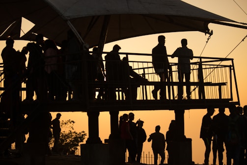 Silhouettes of people gathered on a raised platform against an orange sunset sky. The scene includes a canopy overhead, casting shadows, and people standing, sitting, and engaging in conversation.
