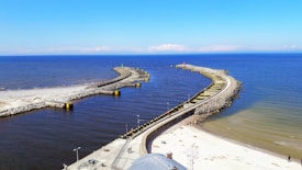 Aerial view of a coastal area with two long, curved breakwaters extending into the sea, creating a protected harbor. The water is a deep blue, contrasting with the sandy beach and rocky structures. The sky is clear with a few clouds visible on the horizon. Some people are walking along the breakwater and beach, while street lamps and small structures are seen alongside the path.