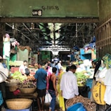 A bustling indoor market with various vendors selling a wide array of fresh produce, including vegetables and fruits. The market is crowded with people engaging in transactions and carrying bags, creating a lively atmosphere.