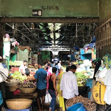 A bustling indoor market with various vendors selling a wide array of fresh produce, including vegetables and fruits. The market is crowded with people engaging in transactions and carrying bags, creating a lively atmosphere.
