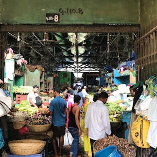 A bustling indoor market with various vendors selling a wide array of fresh produce, including vegetables and fruits. The market is crowded with people engaging in transactions and carrying bags, creating a lively atmosphere.