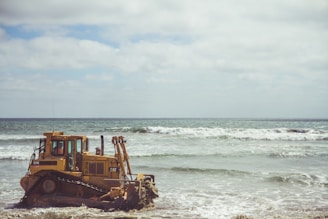 A bulldozer is positioned at the edge of the ocean, partially submerged in water. The sea is slightly wavy with white foam visible on the surface, and the sky above is cloudy yet bright.