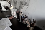 A cluttered artist's workspace featuring a desk with art supplies such as spray cans, brushes, and papers with sketches. There are detailed drawings of trees on the wall, and a decorative raven image is visible on a nearby cabinet.