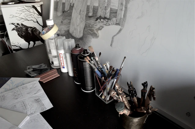 A cozy studio corner with black and white doodles of bugs scattered around a wooden desk filled with art supplies and nature sketches.