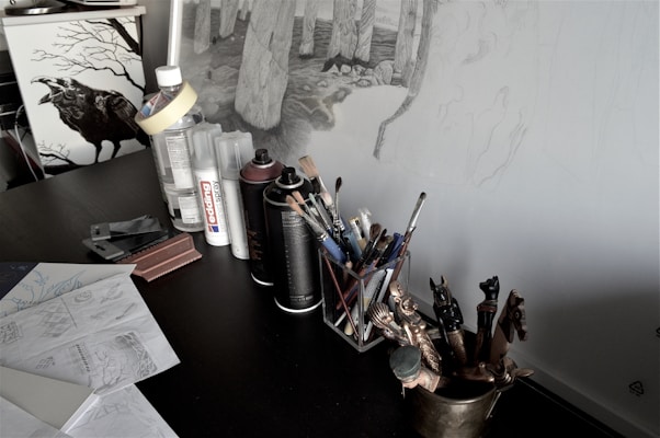 A cluttered artist's workspace featuring a desk with art supplies such as spray cans, brushes, and papers with sketches. There are detailed drawings of trees on the wall, and a decorative raven image is visible on a nearby cabinet.