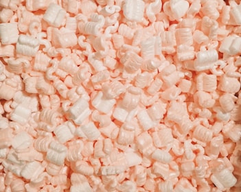 A collection of white and light pink packing peanuts densely packed together, creating a textured surface with curled and cylindrical shapes.