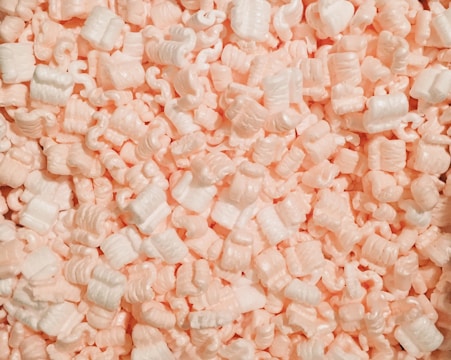 A collection of white and light pink packing peanuts densely packed together, creating a textured surface with curled and cylindrical shapes.