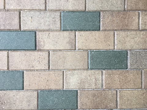 A patterned arrangement of rectangular bricks in alternating shades of light brown and green, with a slightly rough texture and scattered speckles.
