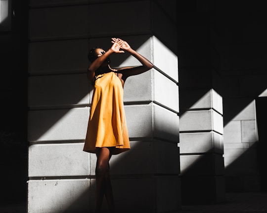 A model wearing a flowing golden-yellow dress standing against a sunlit European street backdrop.