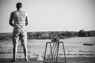 A person is standing on a golf range with their back to the camera, wearing a striped tank top and shorts. A pyramid of golf balls is neatly stacked nearby, and several golf clubs are resting against a stand. The setting is outdoors with a clear sky and flat, expansive terrain.