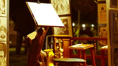 An art setup featuring an easel with a blank paper sheet is spotlighted, surrounded by caricature drawings displayed on wooden stands. Wooden stools and a rustic night ambiance are present.
