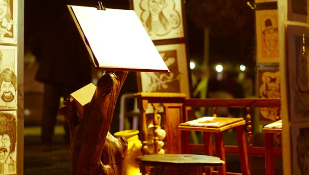 An art setup featuring an easel with a blank paper sheet is spotlighted, surrounded by caricature drawings displayed on wooden stands. Wooden stools and a rustic night ambiance are present.