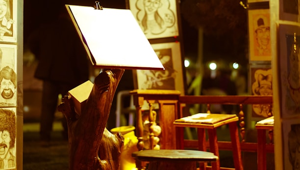 An art setup featuring an easel with a blank paper sheet is spotlighted, surrounded by caricature drawings displayed on wooden stands. Wooden stools and a rustic night ambiance are present.