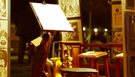 An art setup featuring an easel with a blank paper sheet is spotlighted, surrounded by caricature drawings displayed on wooden stands. Wooden stools and a rustic night ambiance are present.