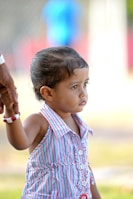 A young child wearing a colorful striped dress is holding onto an adult's hand, suggesting a sense of security. The child's expression appears contemplative as they look into the distance. The background is blurred, emphasizing the child.