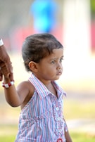 A young child wearing a colorful striped dress is holding onto an adult's hand, suggesting a sense of security. The child's expression appears contemplative as they look into the distance. The background is blurred, emphasizing the child.