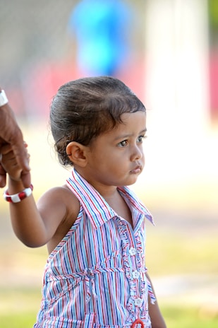 A young child wearing a colorful striped dress is holding onto an adult's hand, suggesting a sense of security. The child's expression appears contemplative as they look into the distance. The background is blurred, emphasizing the child.