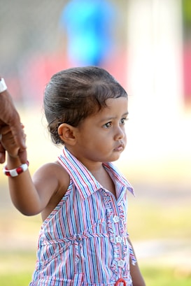 A young child wearing a colorful striped dress is holding onto an adult's hand, suggesting a sense of security. The child's expression appears contemplative as they look into the distance. The background is blurred, emphasizing the child.
