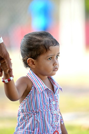 A young child wearing a colorful striped dress is holding onto an adult's hand, suggesting a sense of security. The child's expression appears contemplative as they look into the distance. The background is blurred, emphasizing the child.