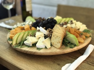 A wooden platter is elegantly arranged with a variety of cheeses, sliced fruits, nuts, crackers, and dried apricots. The presentation is complemented by a glass of white wine nearby, with a soft focus background creating a warm and inviting atmosphere.