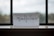 A piece of paper with the word 'mindfulness' written in elegant, flowing cursive sits on a windowsill. The background shows an out-of-focus view through a window, with a blend of muted colors.