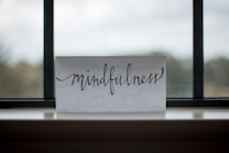 A piece of paper with the word 'mindfulness' written in elegant, flowing cursive sits on a windowsill. The background shows an out-of-focus view through a window, with a blend of muted colors.