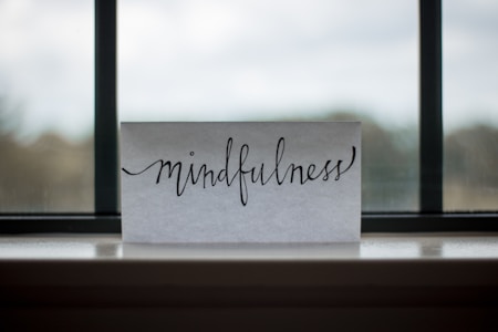 A piece of paper with the word 'mindfulness' written in elegant, flowing cursive sits on a windowsill. The background shows an out-of-focus view through a window, with a blend of muted colors.
