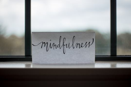 A piece of paper with the word 'mindfulness' written in elegant, flowing cursive sits on a windowsill. The background shows an out-of-focus view through a window, with a blend of muted colors.