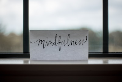 A piece of paper with the word 'mindfulness' written in elegant, flowing cursive sits on a windowsill. The background shows an out-of-focus view through a window, with a blend of muted colors.