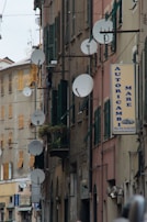 A narrow urban street with closely packed buildings features multiple satellite dishes mounted on the facade. The buildings are characterized by aging exteriors with shutters on the windows. Signs of businesses are visible, including a sign for an auto repair store. The overall appearance suggests a dense, possibly residential area with commercial elements.