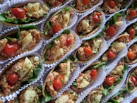 A collection of small, colorful appetizers arranged neatly in paper liners. Each piece is composed of a crispy base topped with a mixture of finely chopped herbs, diced tomatoes, and small chunks of white cheese, all garnished with leafy greens.