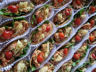 A collection of small, colorful appetizers arranged neatly in paper liners. Each piece is composed of a crispy base topped with a mixture of finely chopped herbs, diced tomatoes, and small chunks of white cheese, all garnished with leafy greens.