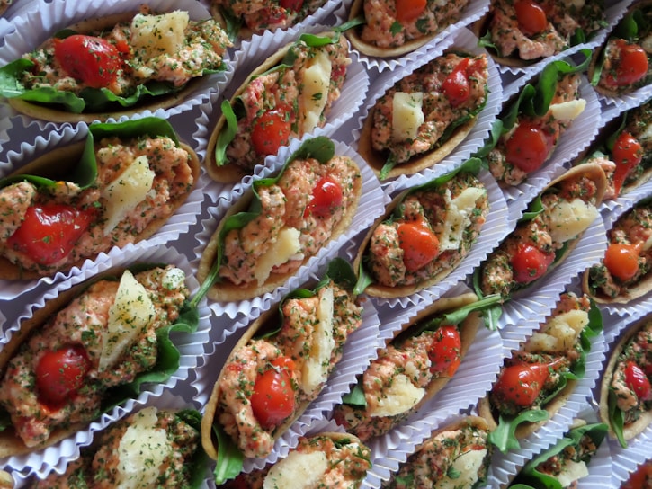 A collection of small, colorful appetizers arranged neatly in paper liners. Each piece is composed of a crispy base topped with a mixture of finely chopped herbs, diced tomatoes, and small chunks of white cheese, all garnished with leafy greens.
