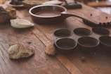 A rustic wooden table is scattered with various kitchen items, including a dark brown frying pan, a metal muffin tin with circular molds, and several rough stones. There is also a white ceramic dish and some indistinct objects in the background, adding to the vintage kitchen ambiance.