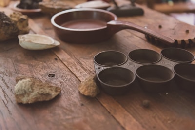 A rustic wooden table is scattered with various kitchen items, including a dark brown frying pan, a metal muffin tin with circular molds, and several rough stones. There is also a white ceramic dish and some indistinct objects in the background, adding to the vintage kitchen ambiance.