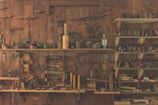 A wooden workshop wall features an array of vintage hand tools neatly organized on shelves and hanging. The tools include hammers, saws, planes, and other carpentry equipment, showcasing a rustic, historical atmosphere.