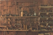 A wooden workshop wall features an array of vintage hand tools neatly organized on shelves and hanging. The tools include hammers, saws, planes, and other carpentry equipment, showcasing a rustic, historical atmosphere.