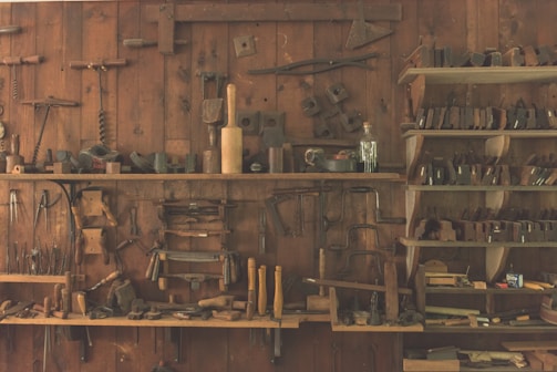 A wooden workshop wall features an array of vintage hand tools neatly organized on shelves and hanging. The tools include hammers, saws, planes, and other carpentry equipment, showcasing a rustic, historical atmosphere.