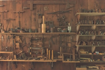 A wooden workshop wall features an array of vintage hand tools neatly organized on shelves and hanging. The tools include hammers, saws, planes, and other carpentry equipment, showcasing a rustic, historical atmosphere.