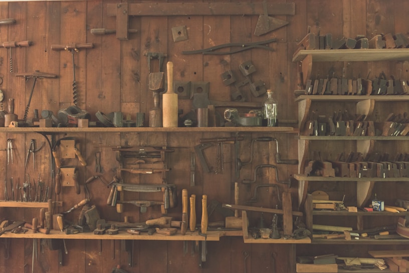 A wooden workshop wall features an array of vintage hand tools neatly organized on shelves and hanging. The tools include hammers, saws, planes, and other carpentry equipment, showcasing a rustic, historical atmosphere.