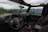 The interior of a Jeep vehicle with a view of the driver's seat, steering wheel, and dashboard is visible. Through the open windows, a scenic landscape with dense evergreen trees and distant snow-capped mountains under a cloudy sky can be seen. A backpack is placed on the passenger seat, and the door panels and control buttons are clearly visible.